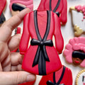Red robe-shaped cookie with black bow held by a hand, surrounded by other decorated cookies.