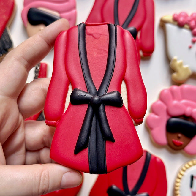 Red robe-shaped cookie with black bow held by a hand, surrounded by other decorated cookies.